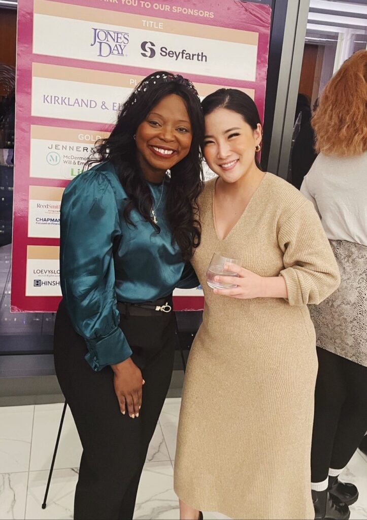 A smiling African-American woman in a teal blouse stands with a smiling Asian-American woman in a camel-colored dress.