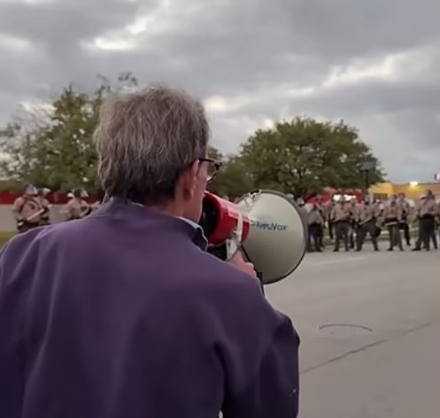 A man in a purple sweatshirt, stands before a line of federal law enforcement, speaking into a bullhorn,