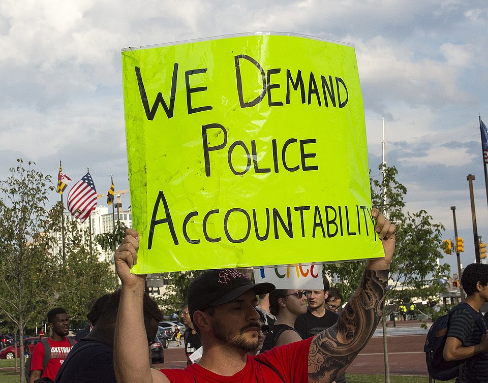 A person holds a sign over their head. The sign reads "We Demand Police Accountability". There is a flag of the United States in the background.